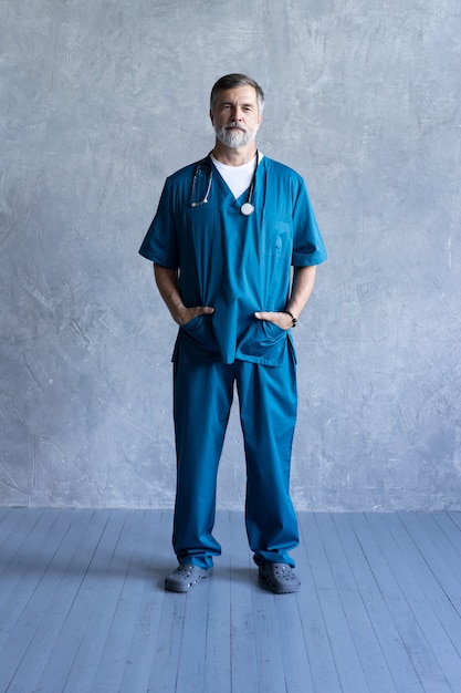 회색 배경에 서서 카메라를 바라보는 전문 성숙한 외과 의사의 전신 초상화.