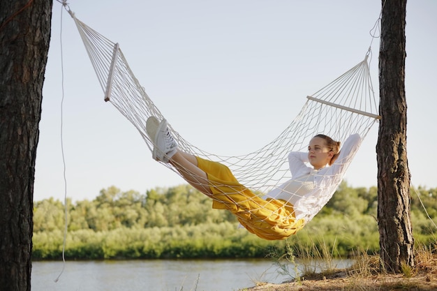 Full length portrait of relaxed calm woman wearing white shirt\
and yellow trousers lying on hammock and looking away enjoying her\
rest in open air posing with river on background