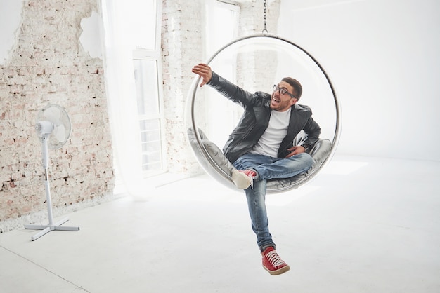 Full-length portrait of relaxed hipster man in casual sitting on hanging chair bullet and smiling on camera isolated over white background.