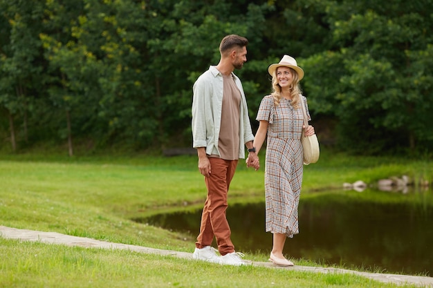 Full length Portrait of romantic adult couple holding hands while walking by lake in rustic countryside scenery