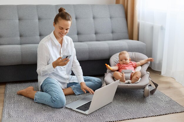 Full length portrait of satisfied woman with bun hairstyle wearing white shirt and jeans young mother looking after baby using smart phone and personal computer