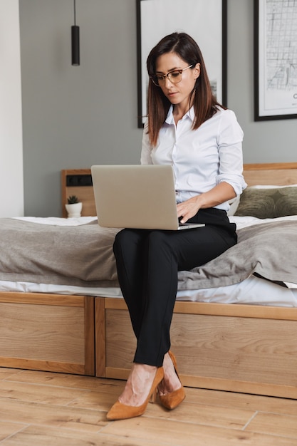 Full length portrait of serious adult businesswoman in formal suit typing on laptop while sitting on bed in apartment