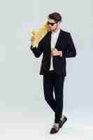 Photo full length portrait of serious young businessman in sunglasses holding yellow skateboard on his sholder over gray wall