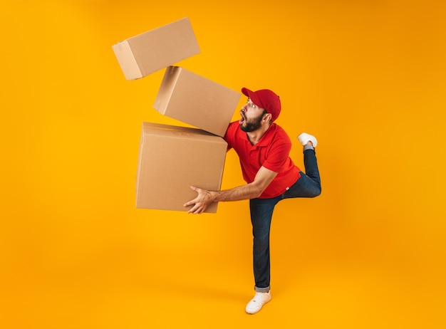 Full length portrait of shocked delivery man in red uniform carrying falling packaging boxes isolated over yellow