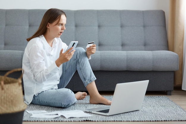 Full length portrait of shocked surprised woman wearing white shirt and jeans sitting on floor with mobile phone and credit card having error with online payment