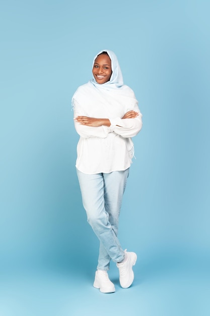 Full length portrait of smiling african american muslim woman\
in headscarf posing with folded arms over blue background