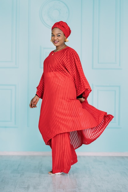 Full length portrait of smiling African woman dressed in stylish traditional ethnic red suit and head scarf, posing on light blue wall studio background.