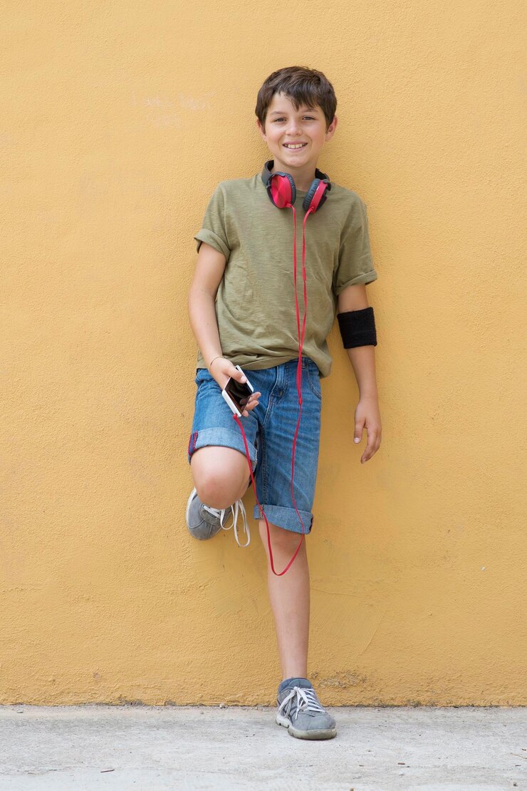 Photo 黄色い壁に立っているスマートフォンとヘッドフォンを持った笑顔の男の子の長さの肖像画