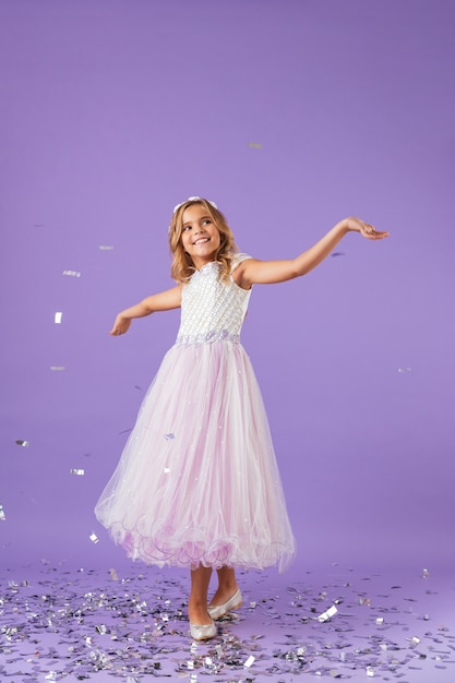 Full length portrait of a smiling cheerful pretty girl dressed in a princess dress isolated over violet wall, dancing