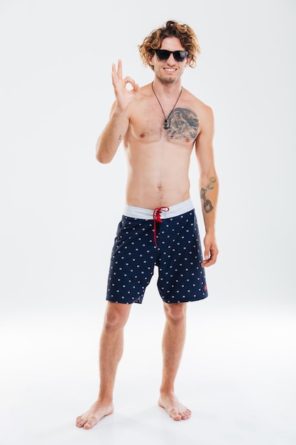 Full length portrait of a smiling curly man in beach shorts showing okay gesture isolated on the white wall