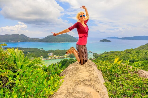 Full length portrait of smiling woman jumping on rock by sea against sky