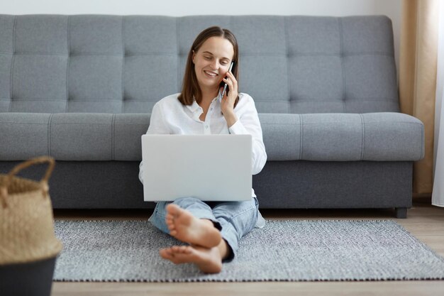 Full length portrait of smiling woman wearing white shirt and jeans sitting on floor near sofa using laptop and talking phone having conversation with friend while working