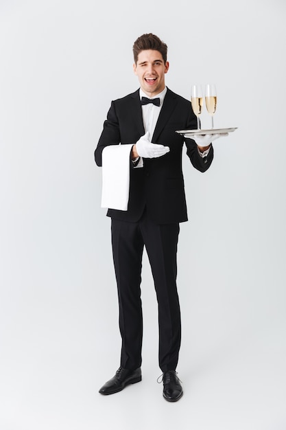 Full length portrait of a smiling young waiter in tuxedo holding tray with two glasses of champagne over white wall