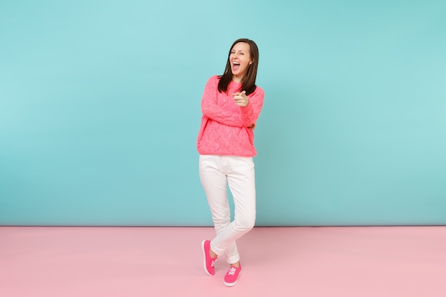Портрет улыбающейся молодой женщины в полный рост в вязаном розовом свитере, позирует в белых штанах