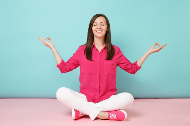 Full length portrait of smiling young woman in rose shirt blouse, white pants sitting on floor 