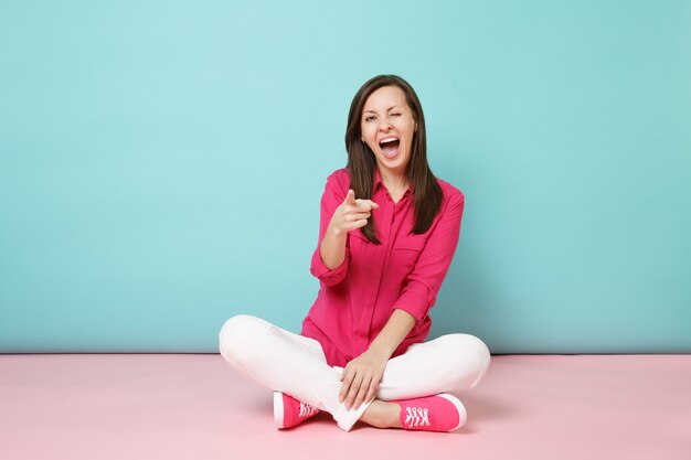 Full length portrait of smiling young woman in rose shirt blouse, white pants sitting on floor 