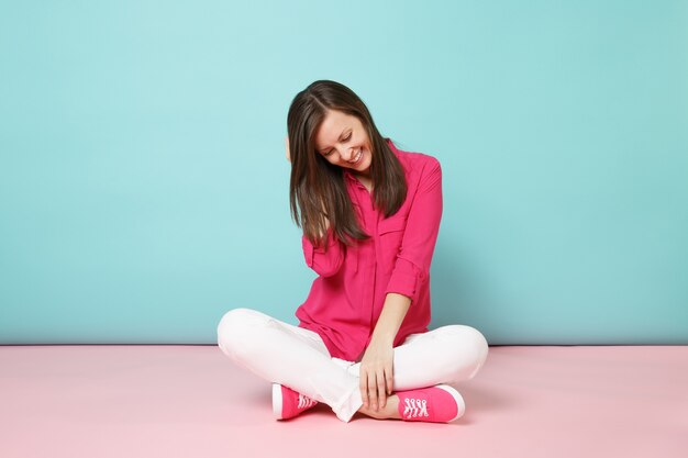 Full length portrait of smiling young woman in rose shirt blouse, white pants sitting on floor 