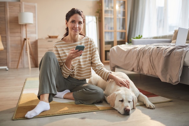 Полный портрет улыбающейся молодой женщины, сидящей на полу, держащей смартфон и собачку в уютном домашнем интерьере, копией пространства
