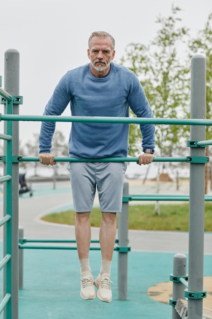 Full length portrait of sportive mature man exercising on parallel bars outdoors and looking at came