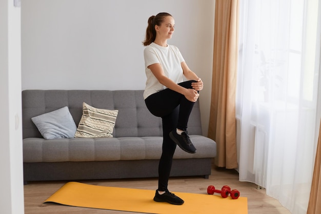 Full length portrait of sporty beautiful woman with ponytail\
wearing black leggins and white t shirt doing sport exercises on\
yoga mat, standing on one leg and doing stretching.