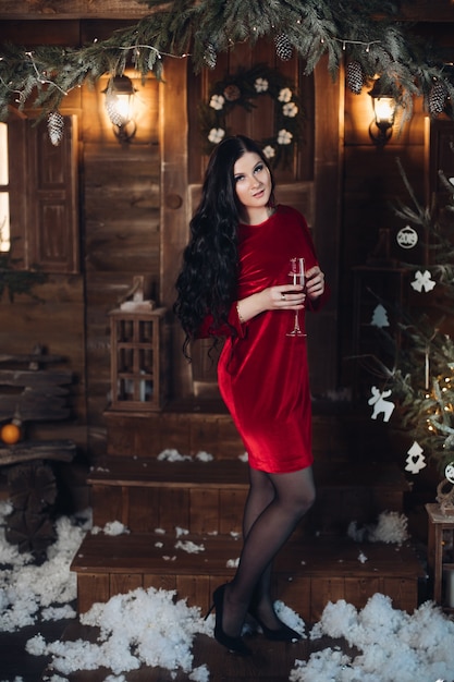 Full length portrait of stunning dark-haired woman in elegant red dress and high heels standing with flute of champagne under snowfall in festive decorated room at Christmas