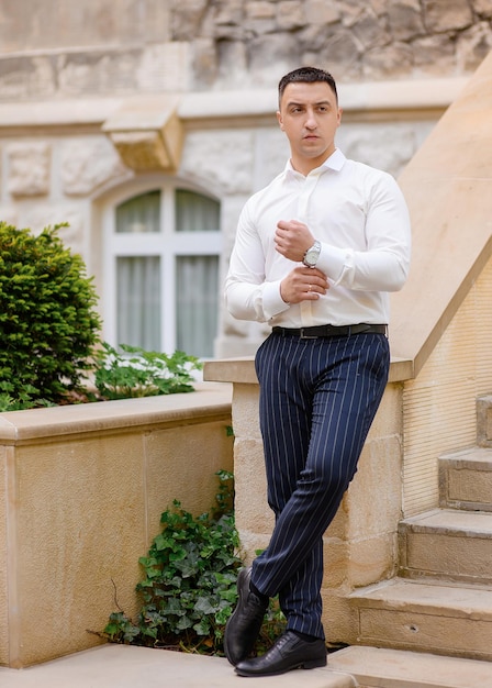 Ritratto integrale dello sposo uomo alla moda che posa lungo vestito con camicie bianche e pantaloni blu scuro uomo bello dello sposo in piedi sulle scale all'aperto guardando avanti matrimonio in attesa della sposa