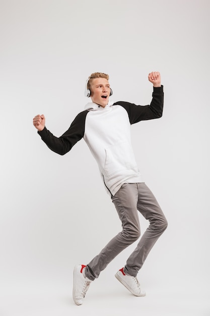 Full length portrait of teenage boy 16-18 years old wearing hoodie and wireless headphones having fun and dancing isolated on white