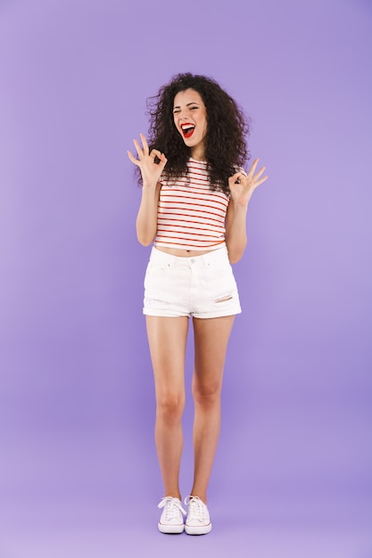 Full length portrait of teenage girl with curly hair in summer wear smiling and showing ok sign