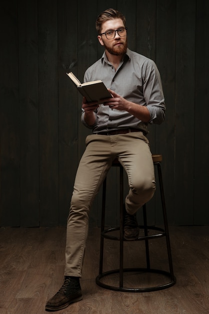 Full length portrait of a thoughtful young man in eyeglasses sitting on chair and reading book isolated on a black wooden surface