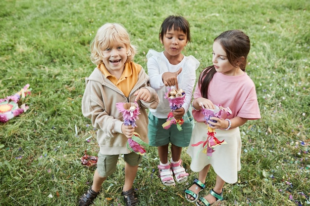 Ritratto a figura intera di tre bambini carini che tengono dolci di pinata durante la festa di compleanno all'aperto