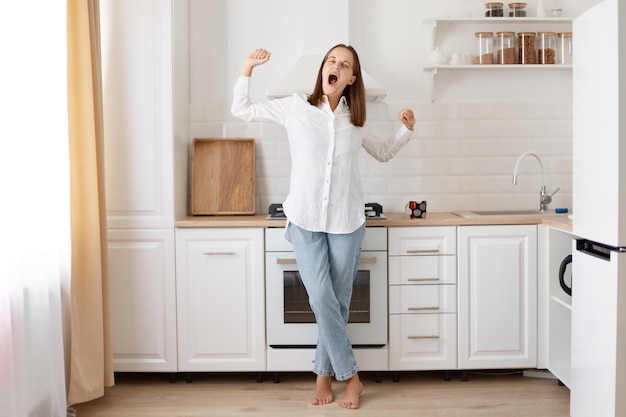 Full length portrait tired lazy woman posing at home in the kitchen, she is stretching arms and yawning, waking up early, needs seep more, wearing white shirt and jeans.