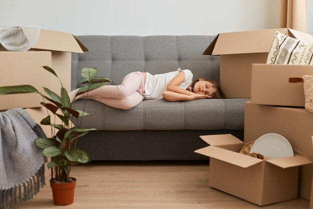 Full length portrait of tired little kid laying on sofa, having nap during relocating and unpacking boxes with belongings, being exhausted and sleepy.