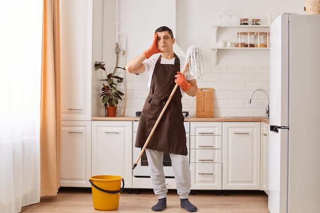 Full length portrait of tired man in rubber gloves and brown apron having a rest from cleaning kitchen floor standing with exhausted facial expression wiping his forehead doing household chores