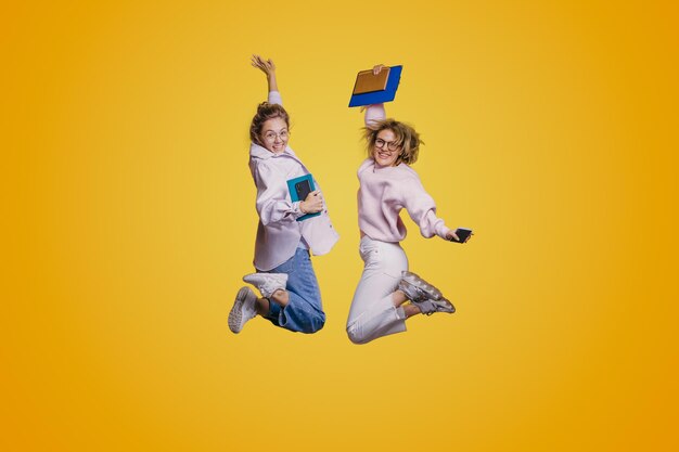 Photo full length portrait of two overjoyed students women in midair holding textbooks and phones isolated on yellow background