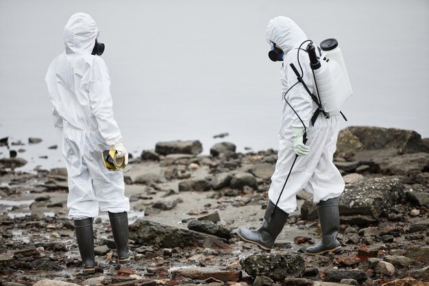 Full length portrait of two workers wearing hazmat suits collecting probe samples by water toxic was...