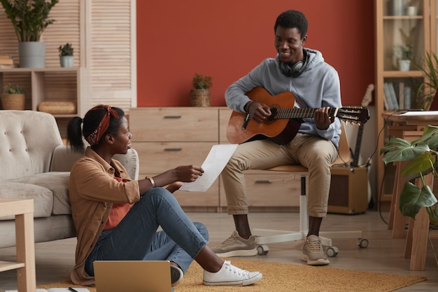 Full length portrait of two young African-American musicians playing guitar and writing music together in home recording studio