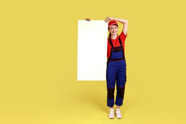 Full length portrait of woman engineer standing and showing big white placard with empty space for advertisement wearing overalls and red cap Indoor studio shot isolated on yellow background