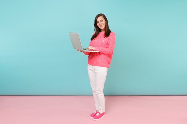 Full length portrait of woman in knitted rose sweater, white pants using laptop pc computer 