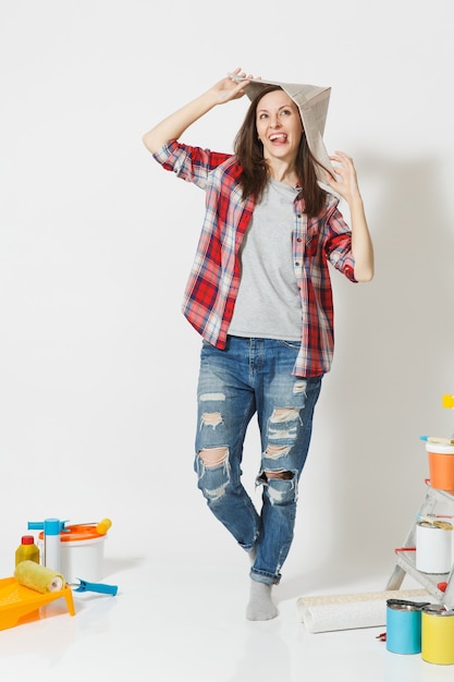 Full length portrait of woman in newspaper hat standing near instruments for renovation apartment home isolated on white background. Wallpaper, accessories for gluing, painting tools. Repair concept.