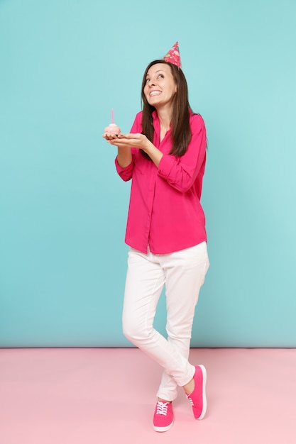 Full length portrait woman in rose shirt blouse, white pants, birthday hat with cupcake posing isolated on bright pink blue pastel wall.