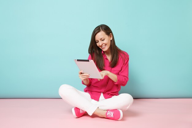 Full length portrait woman in rose shirt blouse, white pants sitting on floor hold tablet pc isolated on bright pink blue pastel wall.