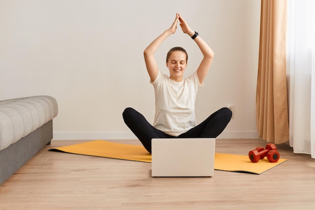 Портрет женщины в полный рост, сидящей на гимнастическом коврике в позе лотоса перед экраном ноутбука, медитирующей, слушающей спокойную музыку, практикующей йогу дома, изучающей асаны из онлайн-класса