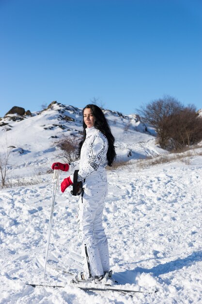 Full Length Portrait of Woman with Long Dark Hair Wearing White Ski Suit and Skis Standing on Snow Covered Mountain and Looking Back Over Shoulder at Camera on Day with Bright Sunshine