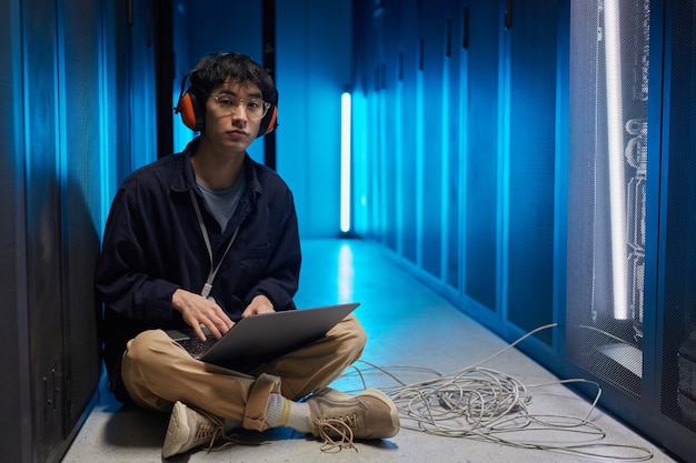 Full length portrait of young Asian man sitting on floor in server room lit by blue light while setting up supercomputer network, copy space