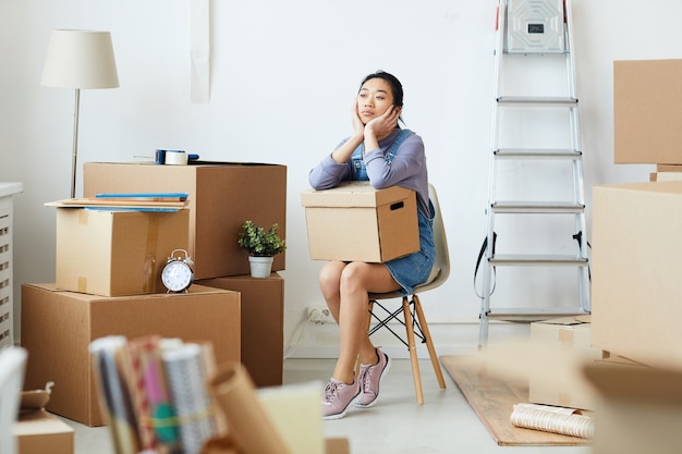 Full length portrait of young Asian woman holding cardboard box sitting on chair in empty room while waiting for moving and relocation crew