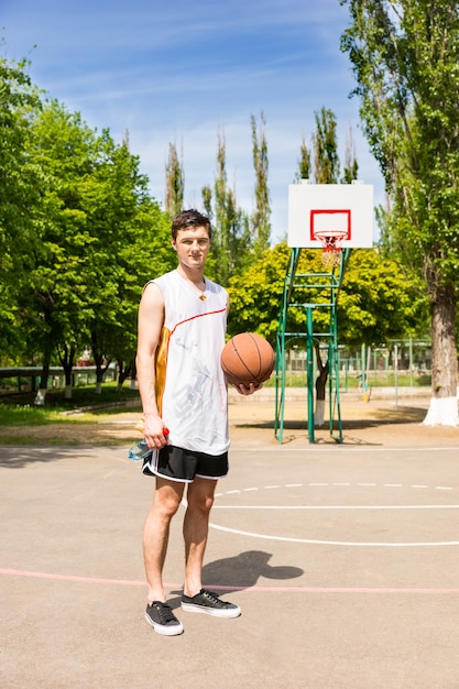 Ritratto integrale di giovane uomo atletico in piedi sul campo da pallacanestro che tiene palla in una luminosa giornata di sole con tabellone e cesto in background