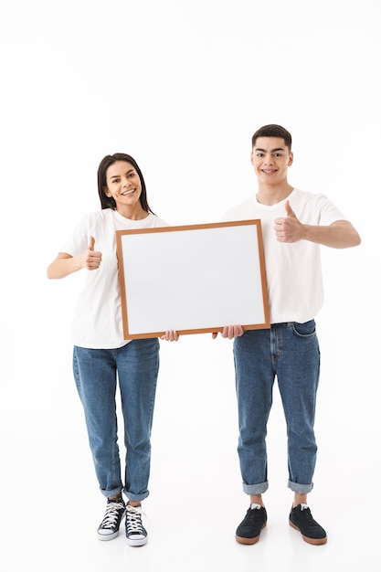 Full length portrait of a young attractive couple standing isolated over white wall, presenting blank board, thumbs up