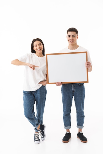 Full length portrait of a young attractive couple standing isolated over white wall, presenting blank board