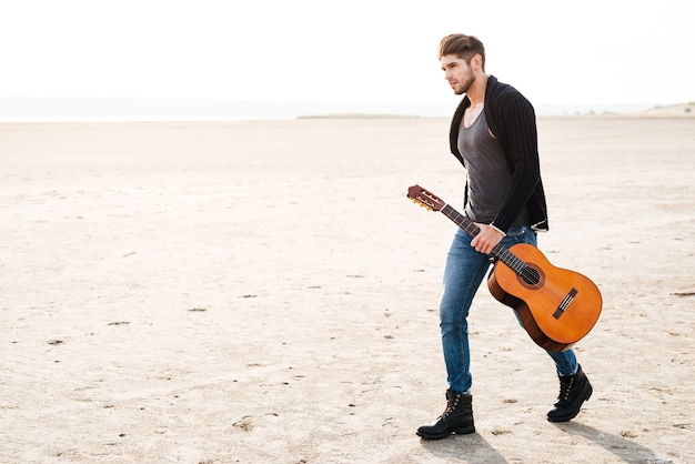 Full length portrait of a young casual man walking across seashore with guitar