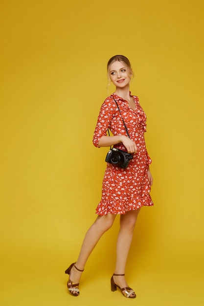 Full-length portrait of a young female traveler in a dress and with a photo camera on the yellow wall. Vacation concept